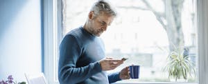 Mature man standing with coffee mug standing in living room looking at smartphone