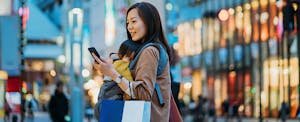 Young Asian mother carrying shopping bag and using smartphone walking in downtown city street shopping with little daughter