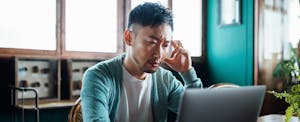 Worried young Asian man with his hand on head, using laptop computer at home, looking concerned and stressed out