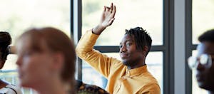 Man sits in classroom with hand raised as if to ask a question