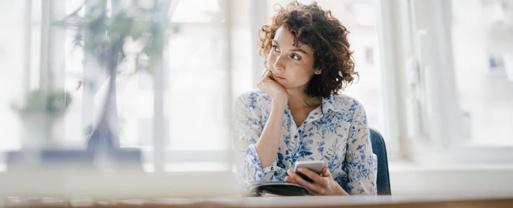 Woman sitting at desk holds her phone and stares into the distance, as though thinking